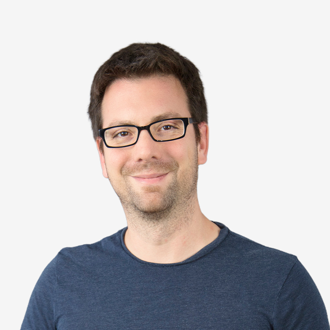 a man smiling in the foreground, wearing glasses and a blue shirt.